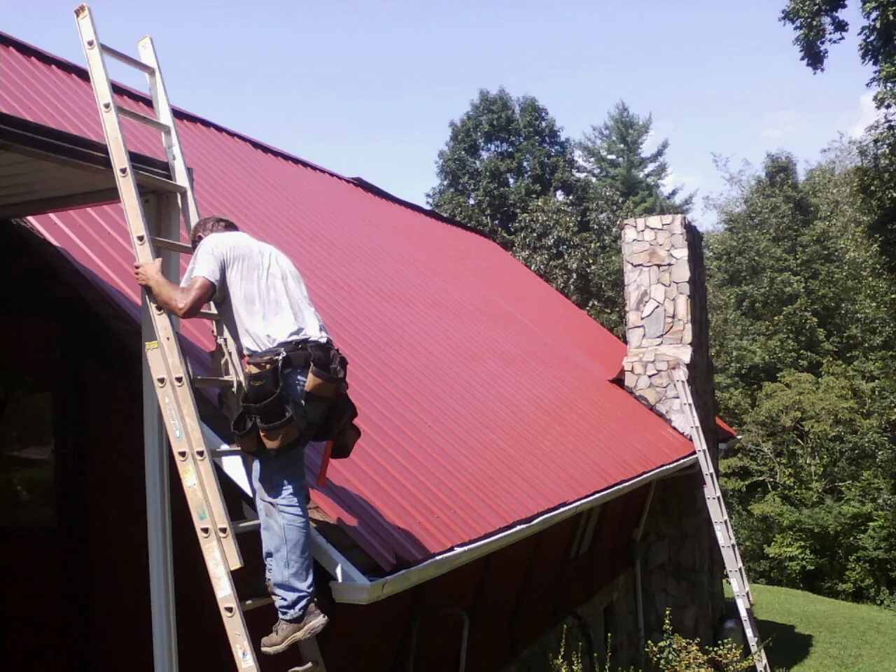 Roofer on a ladder beside a red metal roof and stone chimney — Soffit & Fascia Repair by Roofing Brothers in Donaldson
